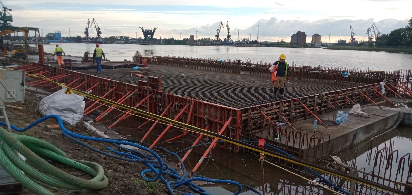 Budowa płyty betonowej z szalunkiem i zbrojeniem na nabrzeżu, widoczni pracownicy w odblaskowych kamizelkach, w tle dźwigi portowe i budynki.
