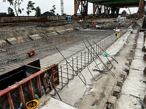 Przygotowanie zbrojenia pod fundament, widoczne metalowe pręty i siatki zbrojeniowe ułożone w wykopie, szalunki drewniane i żółty kask obok, w tle teren budowy z dźwigiem i kontenerami socjalnymi.