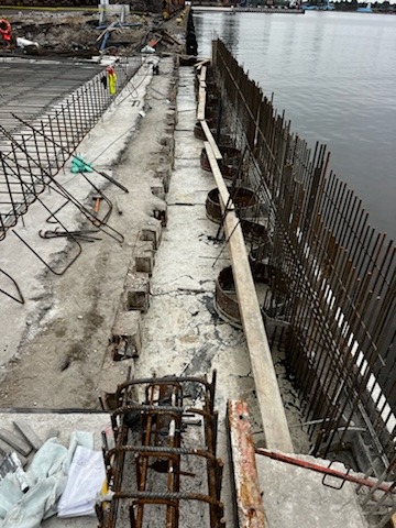 Konstrukcja nabrzeża w trakcie budowy: zbrojenie pionowe i poziome, betonowe elementy konstrukcyjne, widok na wodę i oddalony brzeg.