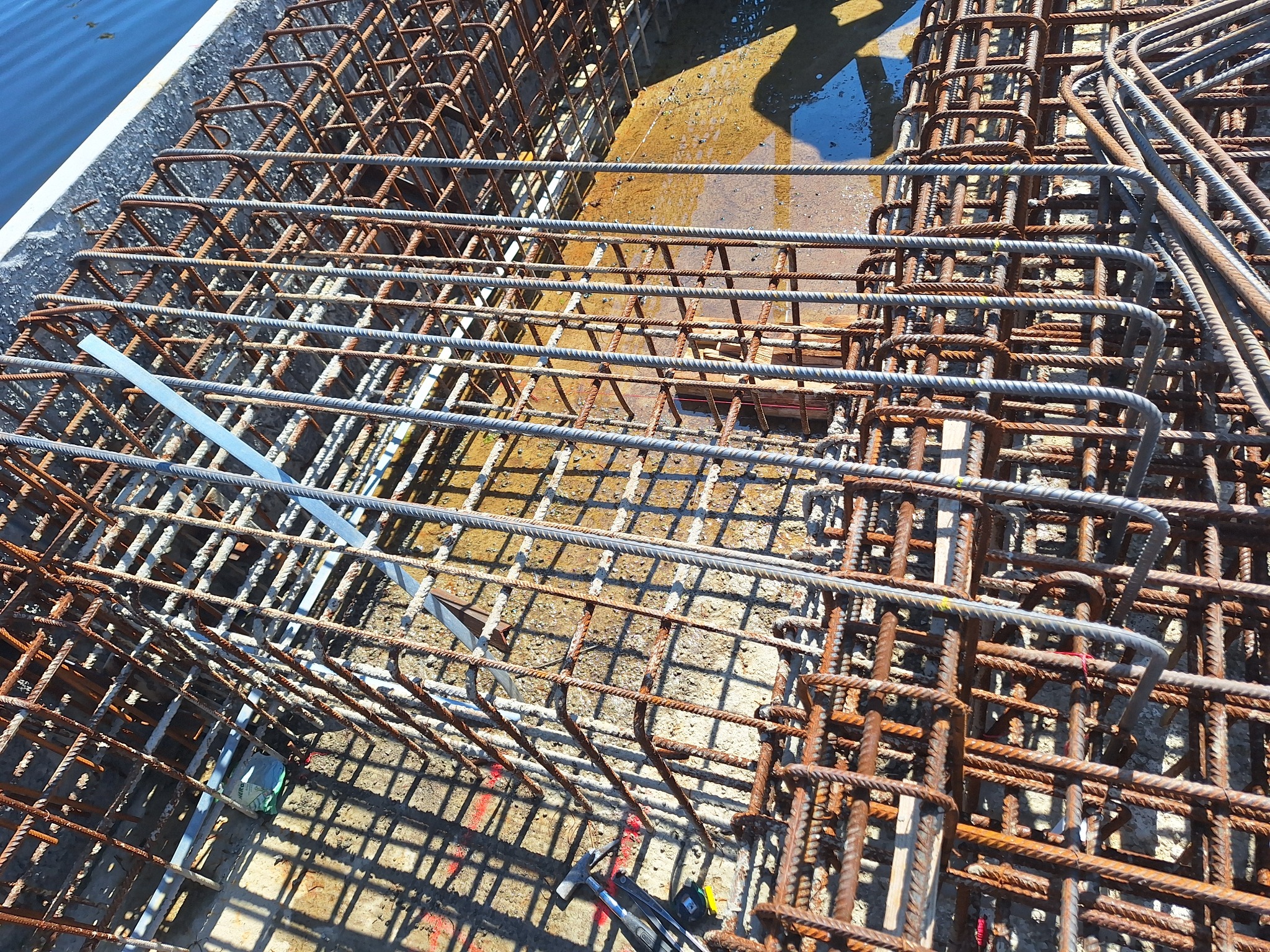 Siatka zbrojeniowa z prętów stalowych o różnym stopniu korozji, ułożona w szalunku, widok z góry z elementami narzędzi w tle.