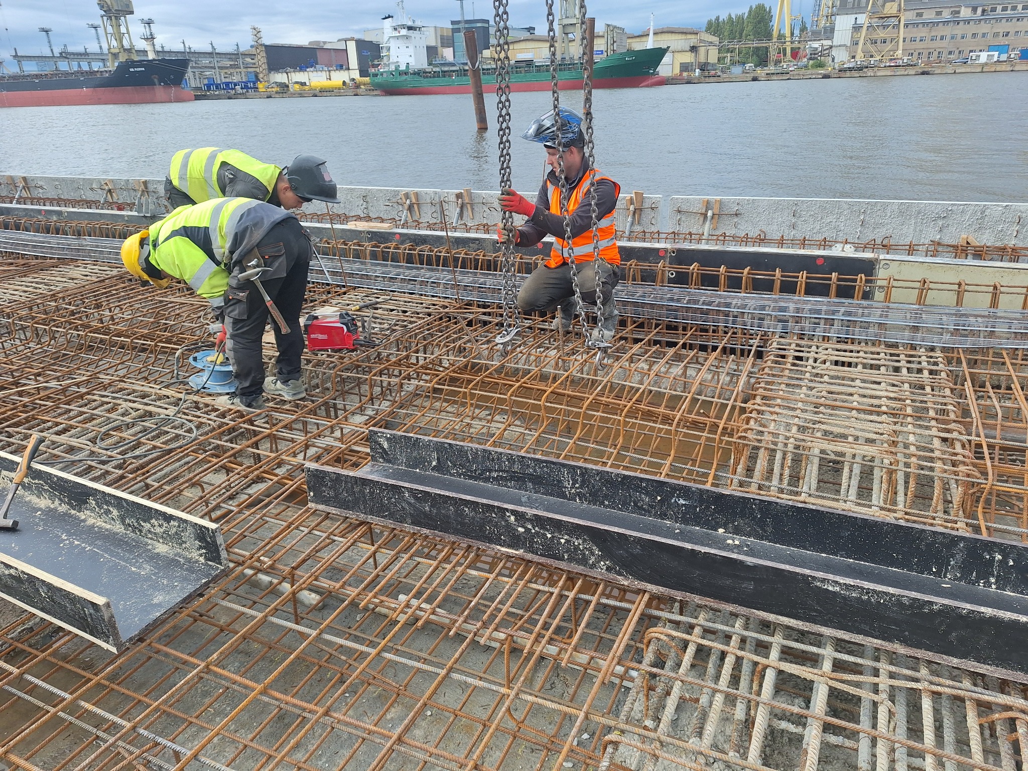 Prace zbrojarskie na budowie nabrzeża: robotnicy w odblaskowych kamizelkach i kaskach układają stalowe pręty zbrojeniowe i czarne kształtowniki w siatkę, w tle widoczny port i statki.