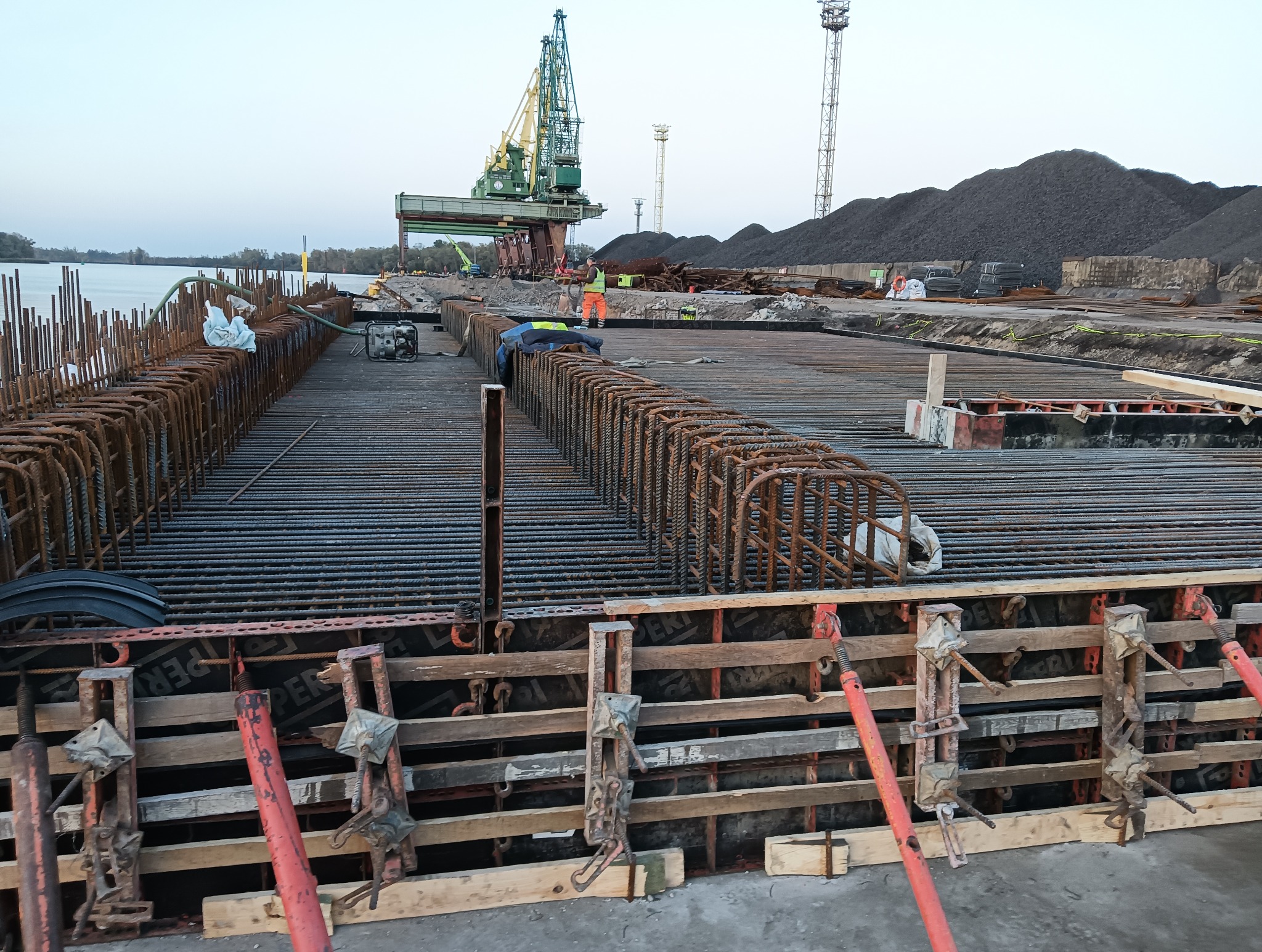 Rozległe zbrojenie płyty fundamentowej z widoczną konstrukcją szalunkową, pracownikami w tle i żurawiem portowym w oddali.