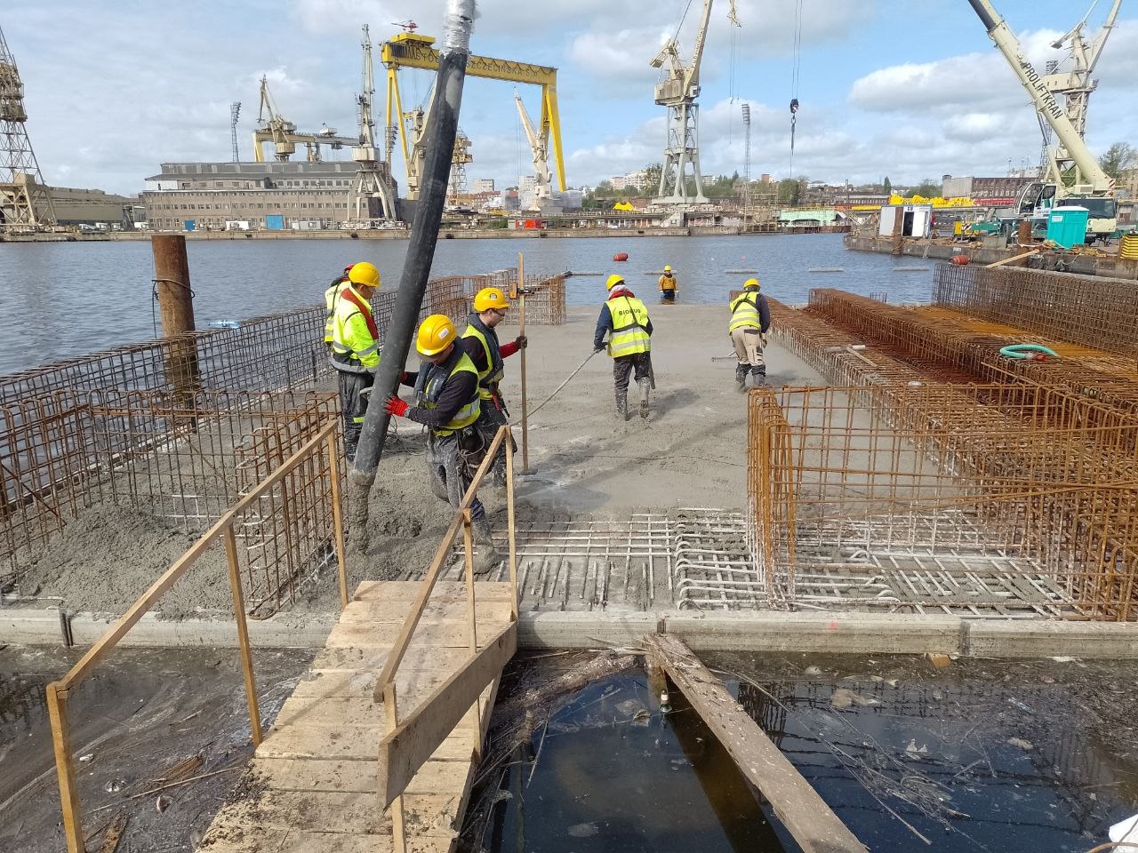 Wylewanie betonu na platformę zbrojoną siatką stalową, pracownicy w żółtych kaskach i kamizelkach, w tle dźwigi portowe i budynki stoczni.