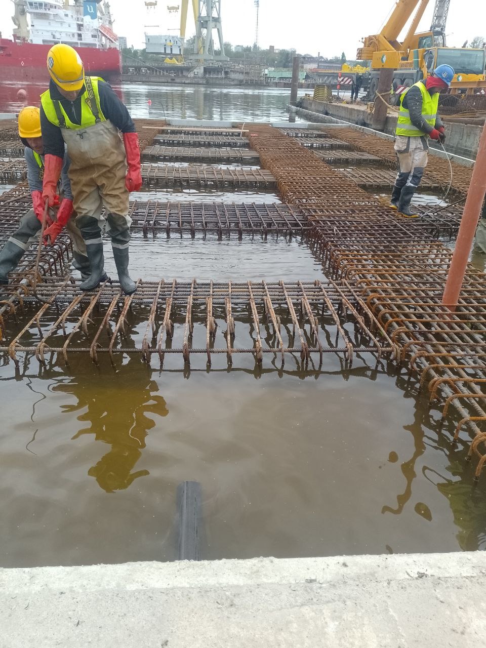 Pracownicy w kombinezonach i kaskach montują zbrojenie w szalunku wypełnionym wodą, w tle widoczny port z dźwigami i statkiem.