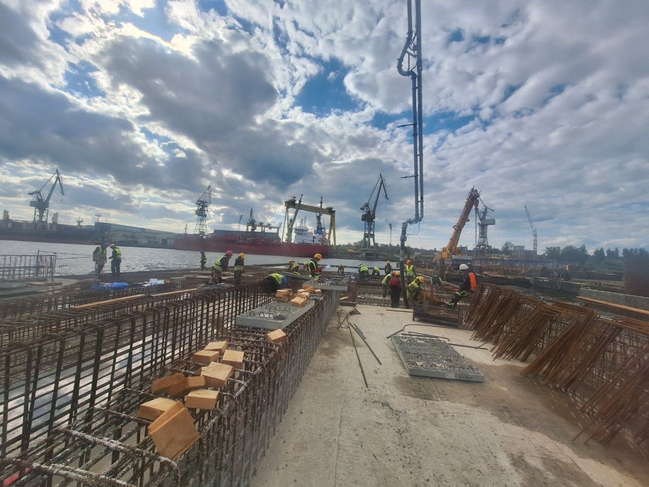Zbrojenie konstrukcji betonowej na nabrzeżu portowym, widoczna ekipa budowlana w kaskach i kamizelkach, pompy do betonu i dźwigi portowe w tle pod zachmurzonym niebem.