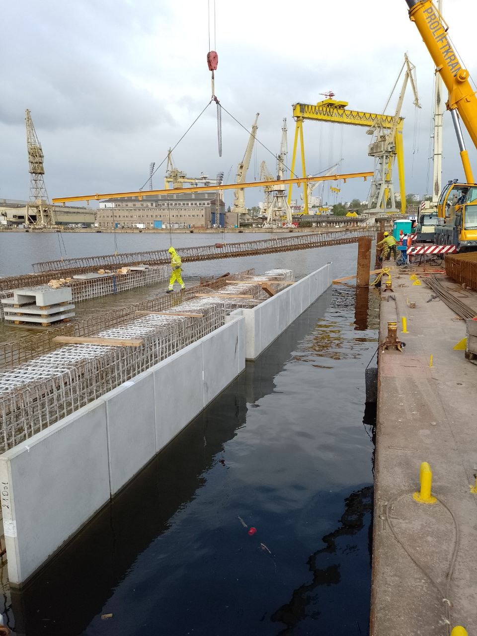 Montaż prefabrykowanych elementów betonowych zbrojonych stalą w środowisku wodnym, widoczne dźwigi portowe w tle.