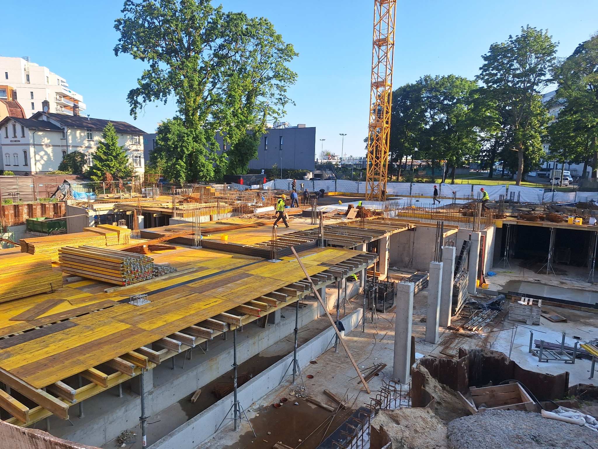 Żółte szalunki i zbrojenia na budowie budynku żelbetowego w słoneczny dzień. W tle drzewa i budynki mieszkalne, a w oddali żółty żuraw budowlany.
