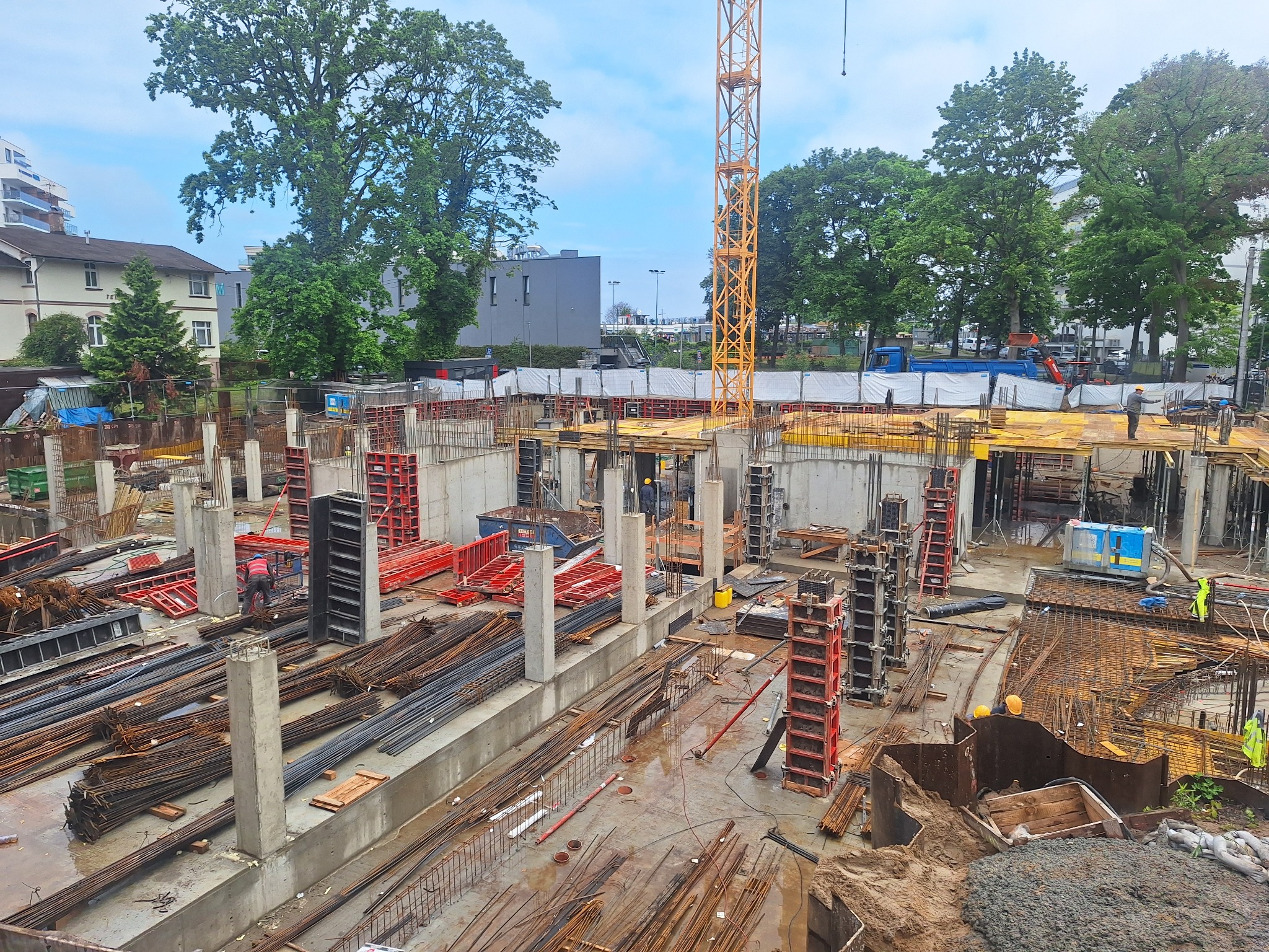 Budowa budynku żelbetowego w toku: widok z góry na zbrojenie, betonowe słupy i szalunki, z żurawiem w tle. Praca wre na placu budowy.