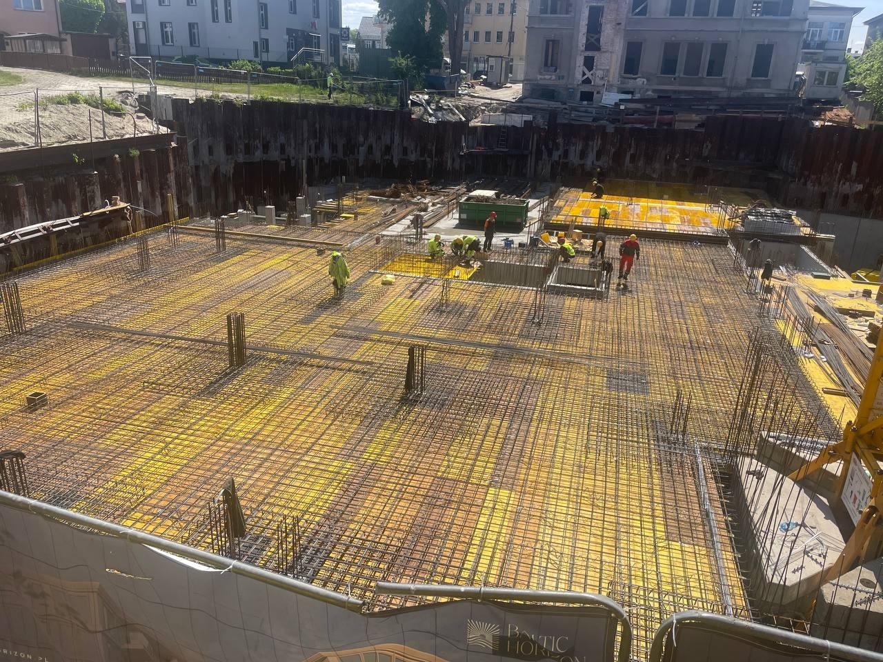 Zbrojenie fundamentu z żółtą folią, widok z góry na plac budowy z pracownikami w odblaskowych ubraniach. Metalowe ścianki oporowe.