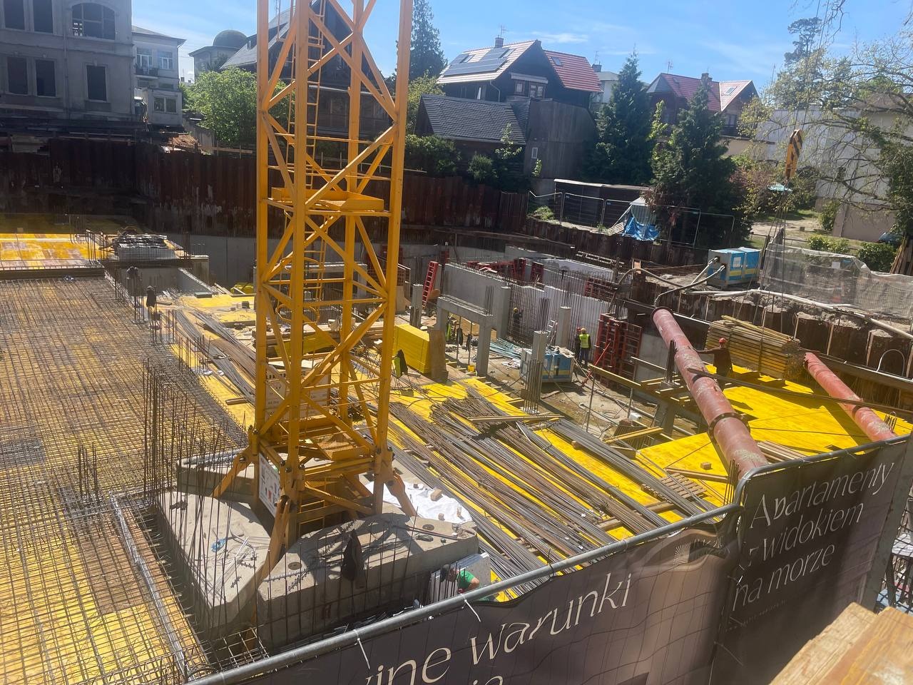 Budowa apartamentów: żółte szalunki, stalowe zbrojenia, betonowe ściany i żuraw na tle zieleni i budynków. Widok z góry na plac budowy.