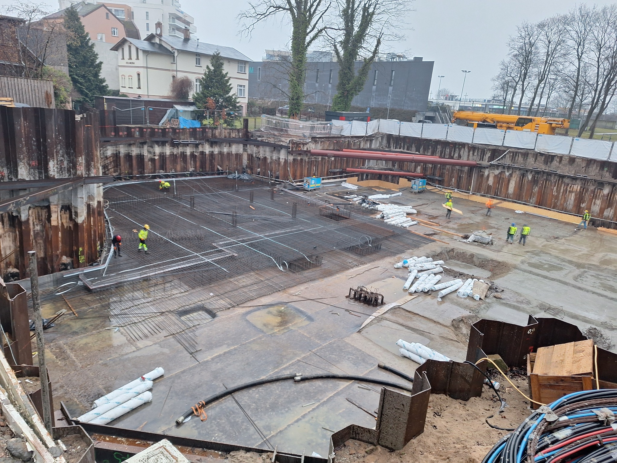 Budowa fundamentów: zbrojenie, stalowe ściany oporowe, robotnicy w żółtych kamizelkach. W tle budynki mieszkalne i drzewa. Widok z góry na plac budowy.