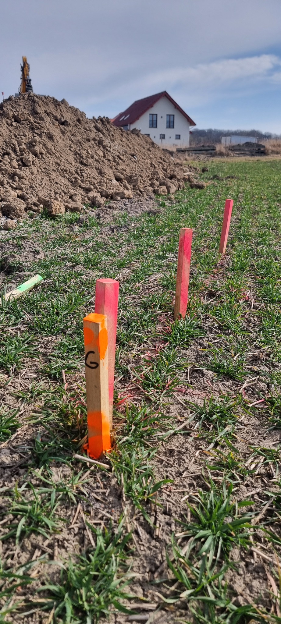 Tyczenie działki: drewniane paliki z oznaczeniami w pomarańczowo-różowym kolorze, w tle kopiec ziemi i budynek. Usługa geodezyjna w trakcie realizacji.