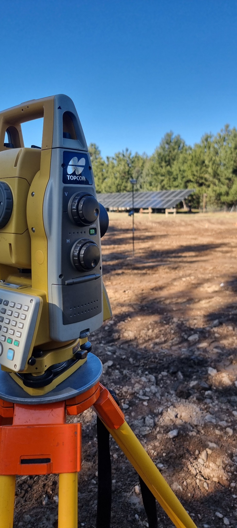 Żółto-szary teodolit Topcon na trójnogu, ustawiony na placu budowy z panelami słonecznymi w tle, pod błękitnym niebem.