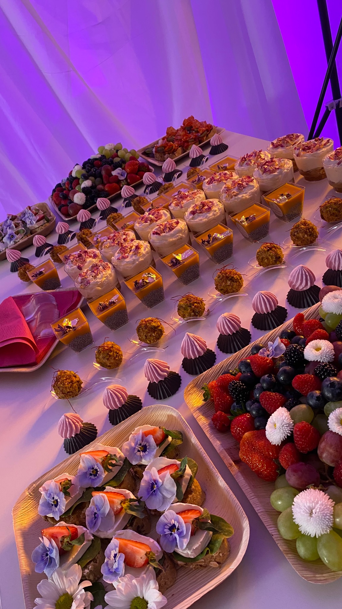 Stół z różnorodnymi przekąskami: mini kanapeczki z kwiatami, desery w szklanych naczyniach, owoce na drewnianych tacach, wszystko oświetlone fioletowym światłem.