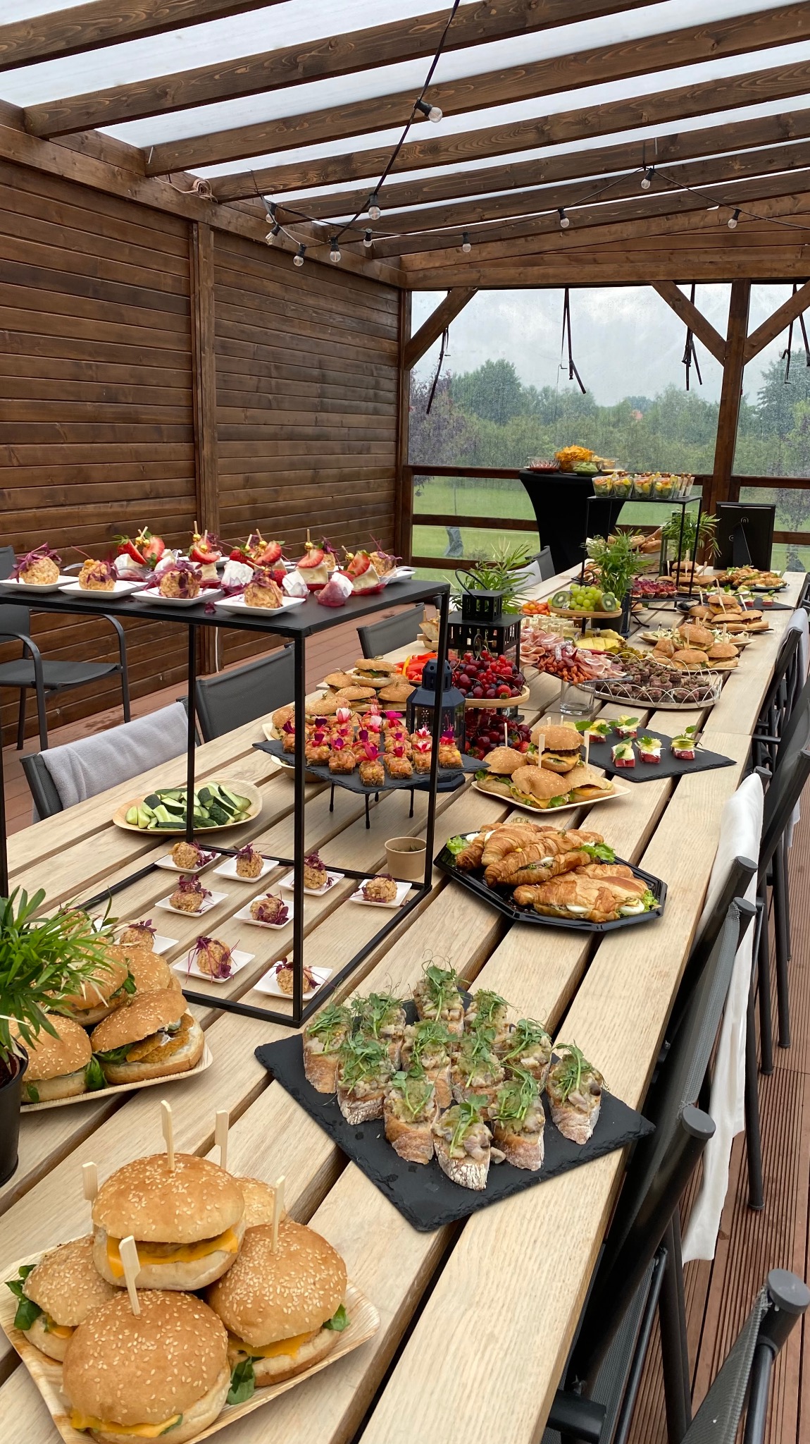Stół cateringowy na zewnątrz pod drewnianą pergolą, pełen różnorodnych przekąsek: mini burgery z wykałaczkami, kanapki z mikrogreens, croissanty, owoce i inne finger food, przygotowany na przyjęcie.