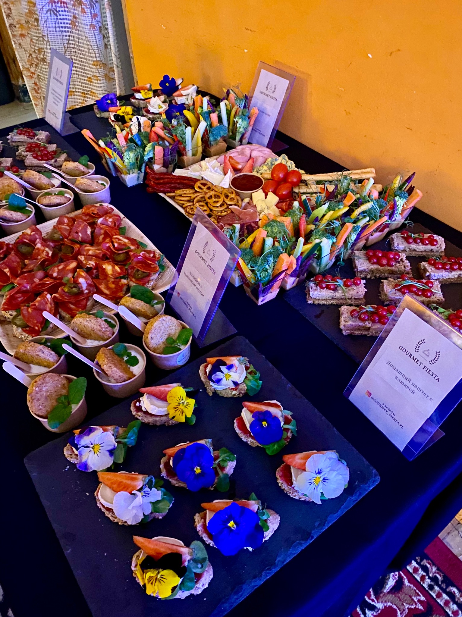 Stół cateringowy z różnorodnymi przekąskami: kanapeczki z salami i jalapeno, mini-deserki w papierowych kubeczkach, warzywa w papierowych foremkach, krakersy z szynką i serem, oraz kanapki...