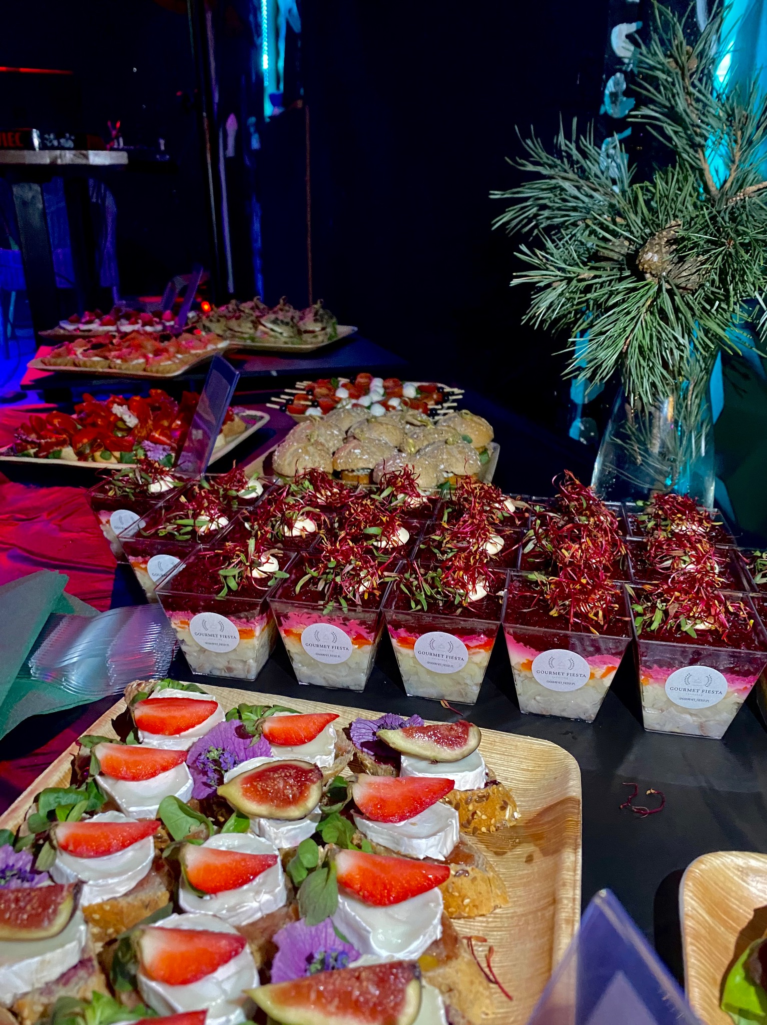 Dekoracyjnie ułożone przekąski i sałatki w słoiczkach na stole podczas eventu, w tle gałązka świerku w wazonie i ciemne tło.