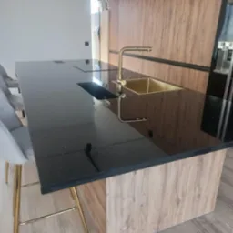 Czarny, błyszczący blat z brokatowym wzorem, z wbudowanym złotym zlewem i baterią, na tle drewnianej zabudowy kuchennej oraz krzesła barowe z złotymi nogami.