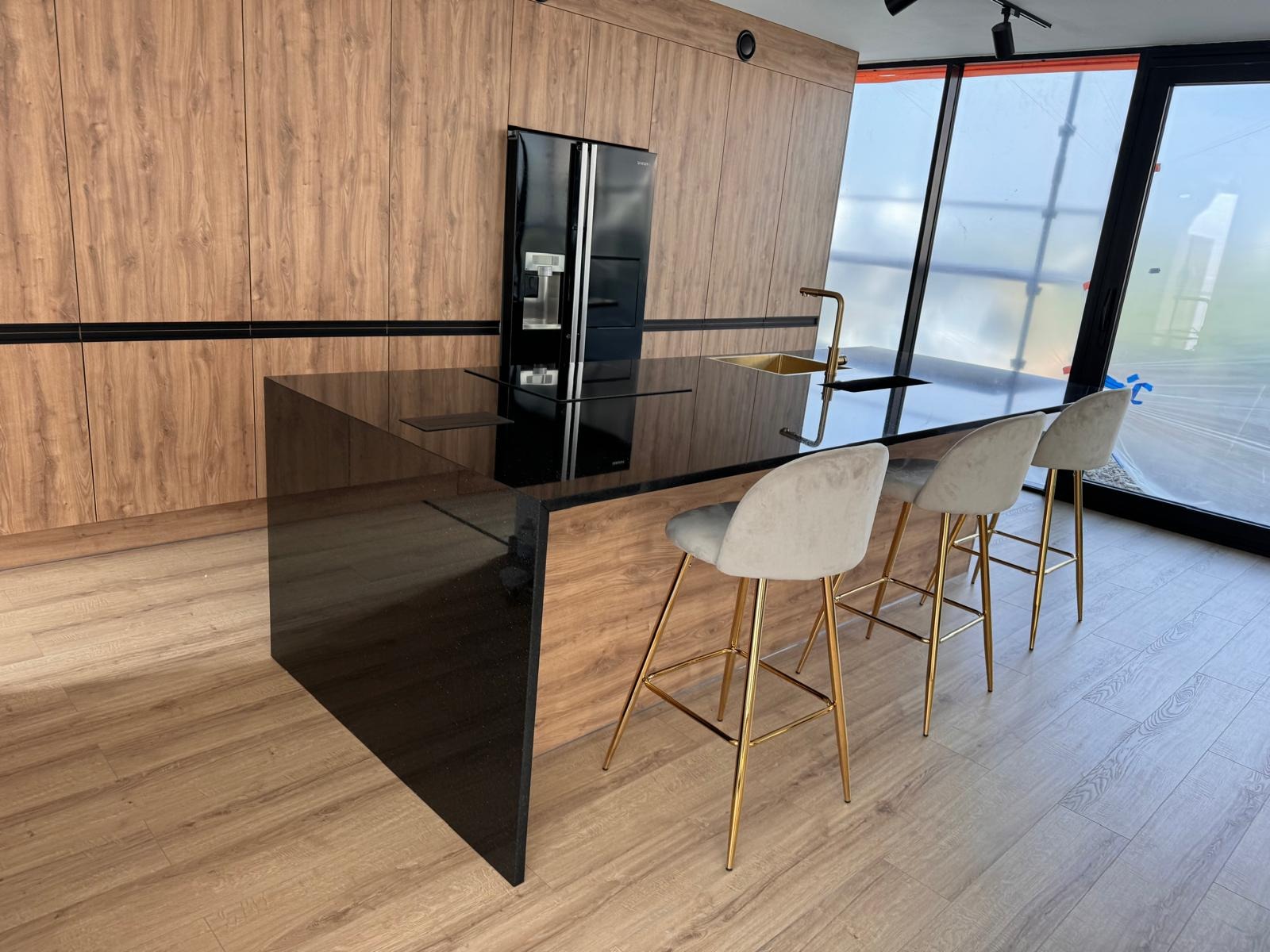 Nowoczesna kuchnia z czarnym blatem z kamienia, złotą armaturą i krzesłami barowymi na złotych nogach, widok na okno w tle.