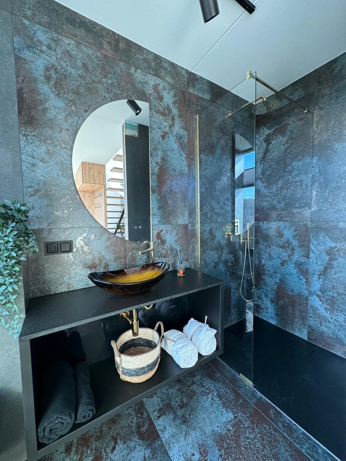 Nowoczesna łazienka z płytkami imitującymi rdzę, okrągłym lustrem, szklaną umywalką i kabiną prysznicową typu walk-in z elementami w kolorze złotym.