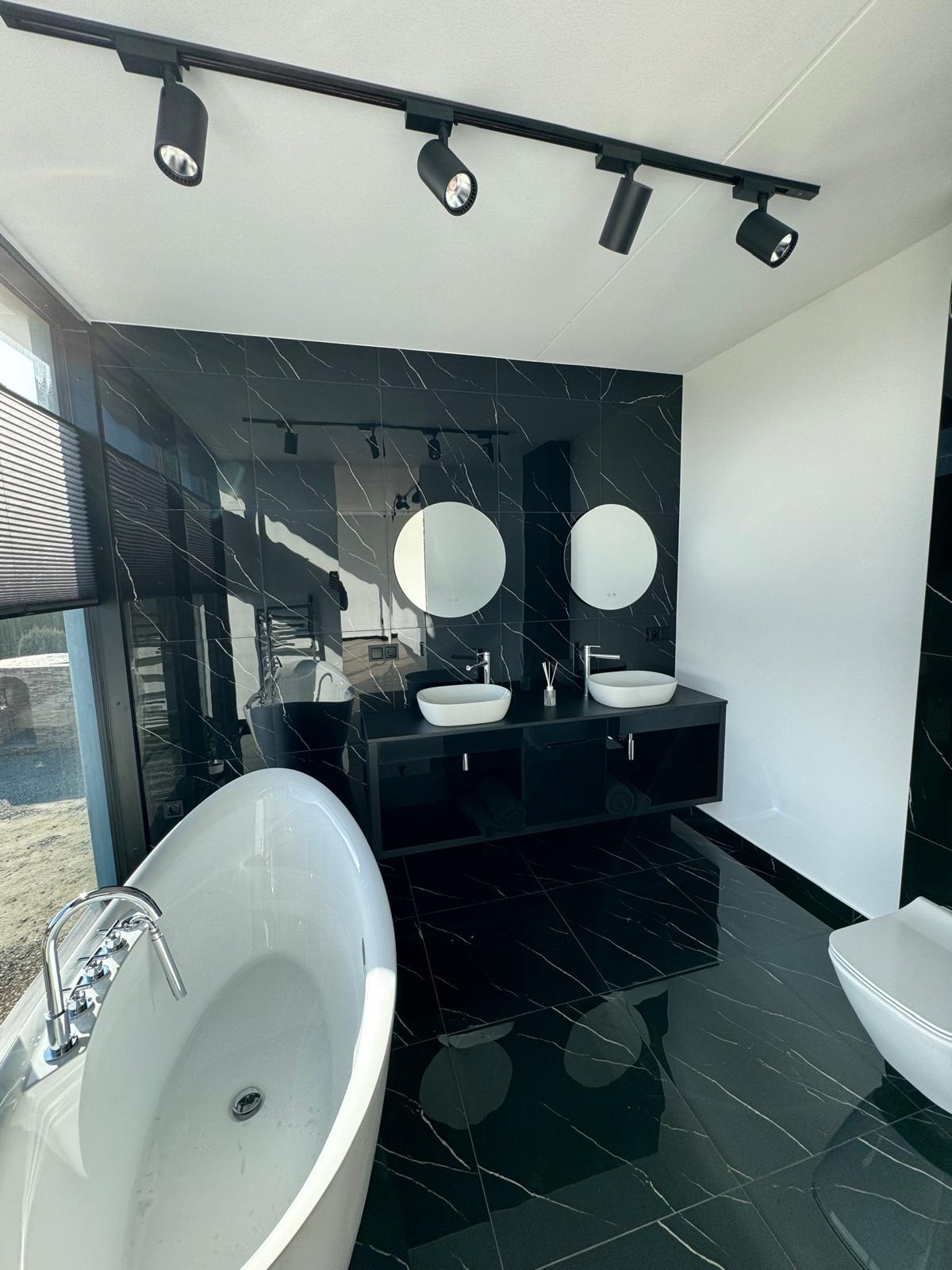Nowoczesna łazienka z czarnymi płytkami imitującymi marmur, owalna wanna, podwójna umywalka z czarną szafką, punktowe oświetlenie sufitowe i duże okno z żaluzjami.