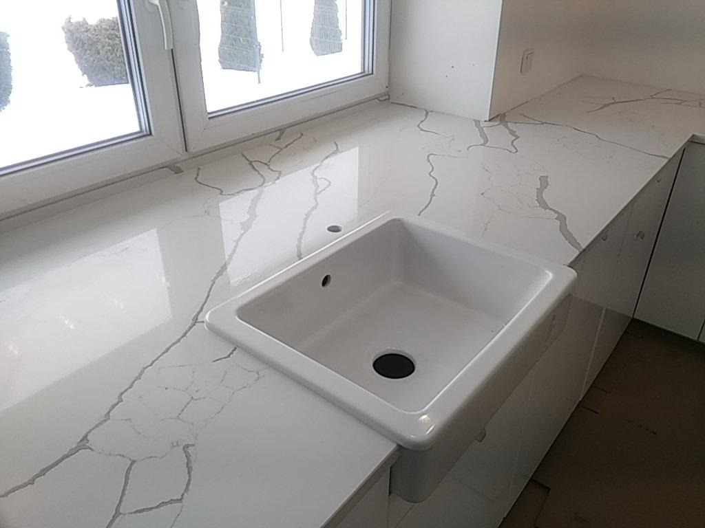 Biały blat kamienny z szarymi żyłami, zintegrowany z kwadratowym zlewem ceramicznym pod oknem z widokiem na zaśnieżony ogród.