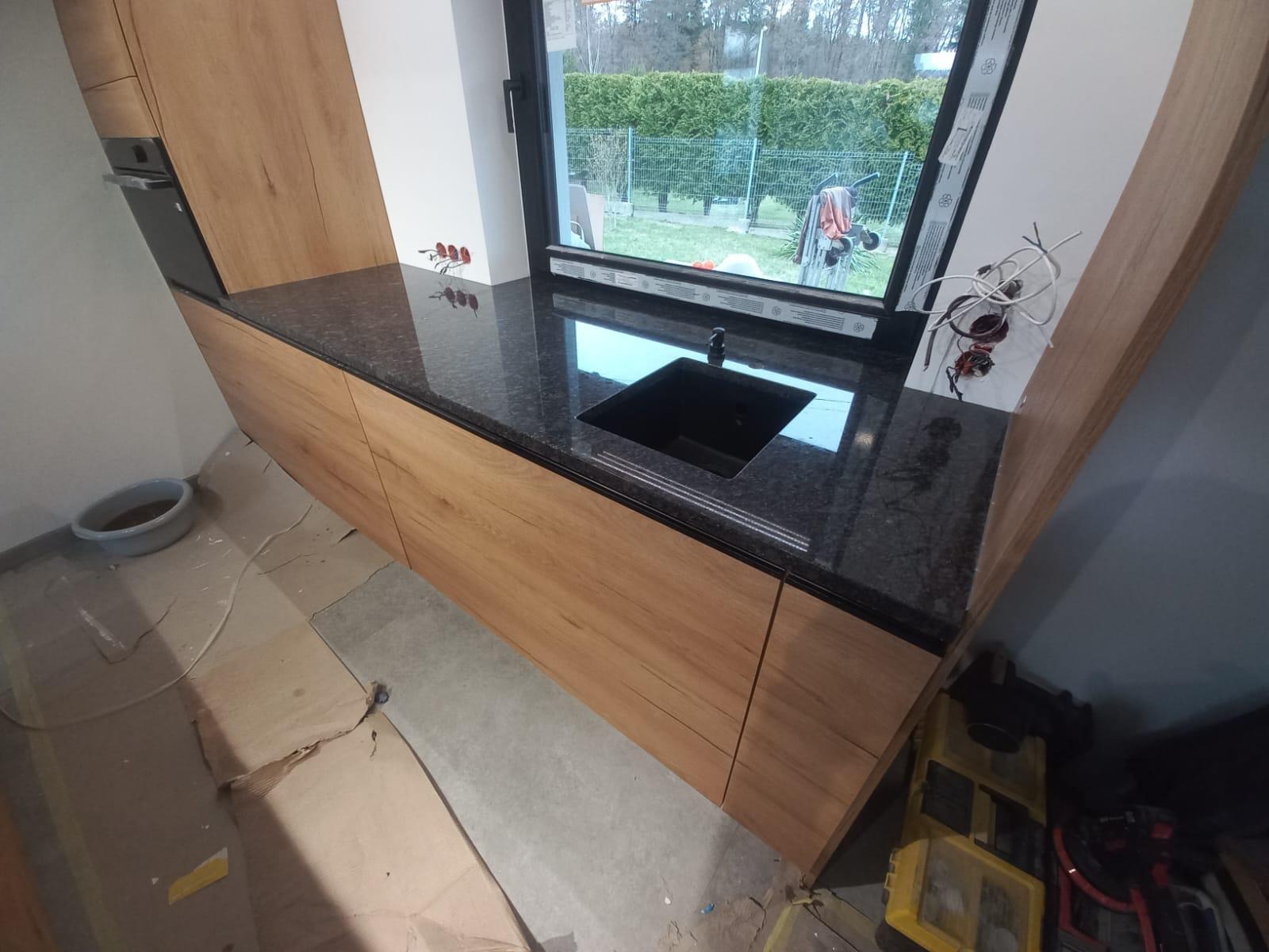 Nowoczesna kuchnia z drewnianymi szafkami i czarnym, błyszczącym blatem z wbudowanym zlewem, widoczne niedokończone podłączenia elektryczne przy oknie i zabezpieczona podłoga.