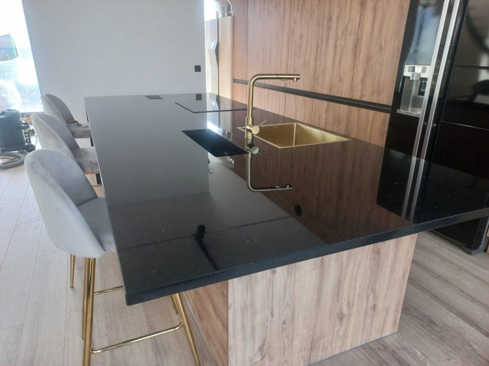 Czarny, błyszczący blat z brokatowym wzorem, z wbudowanym złotym zlewem i baterią, na tle drewnianej zabudowy kuchennej oraz krzesła barowe z złotymi nogami.