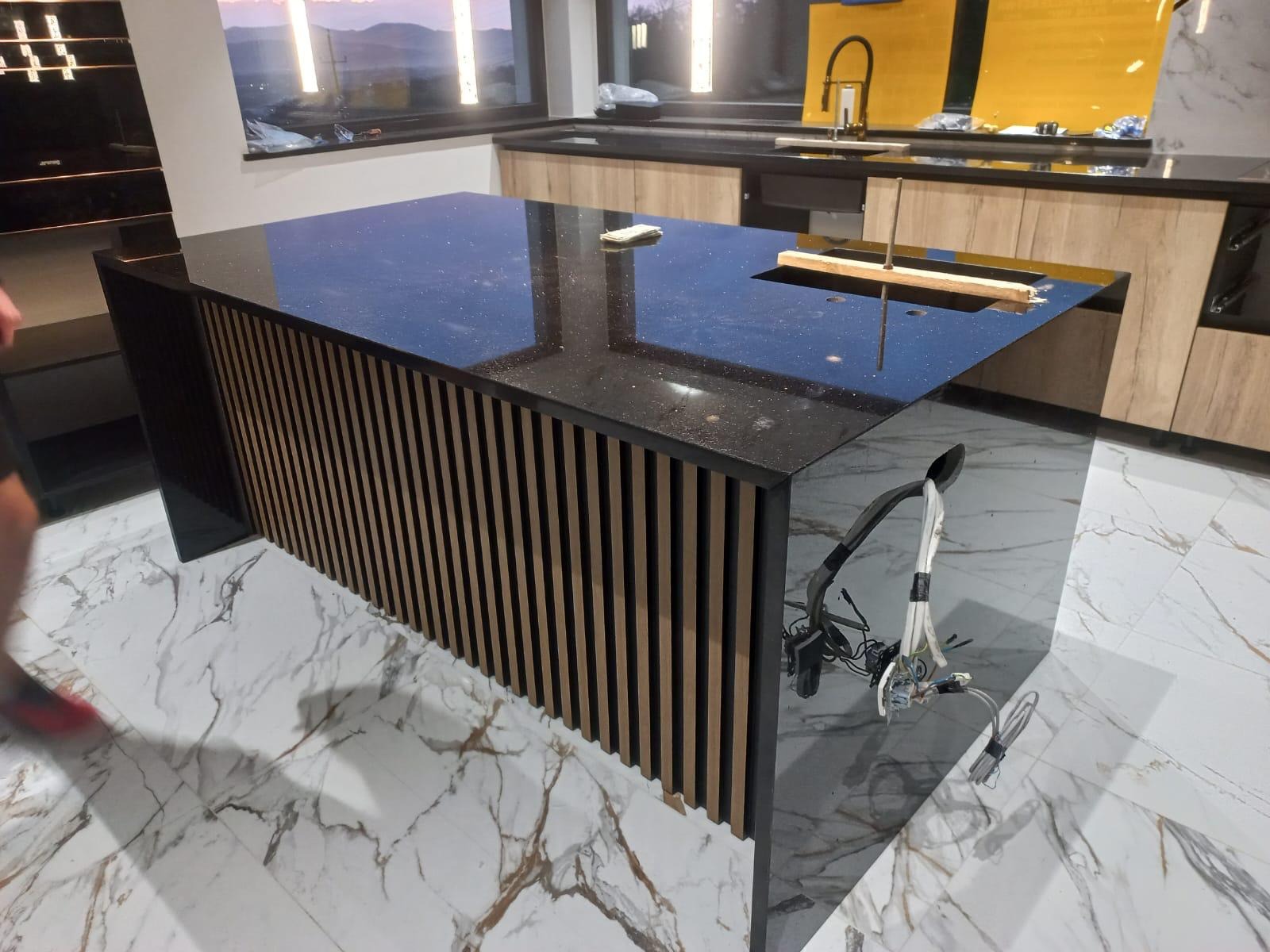Wyspa kuchenna z czarnym blatem z ciemnego konglomeratu z połyskiem, obudowana drewnianymi lamelami, w trakcie montażu oświetlenia, na podłodze marmurowe płytki.