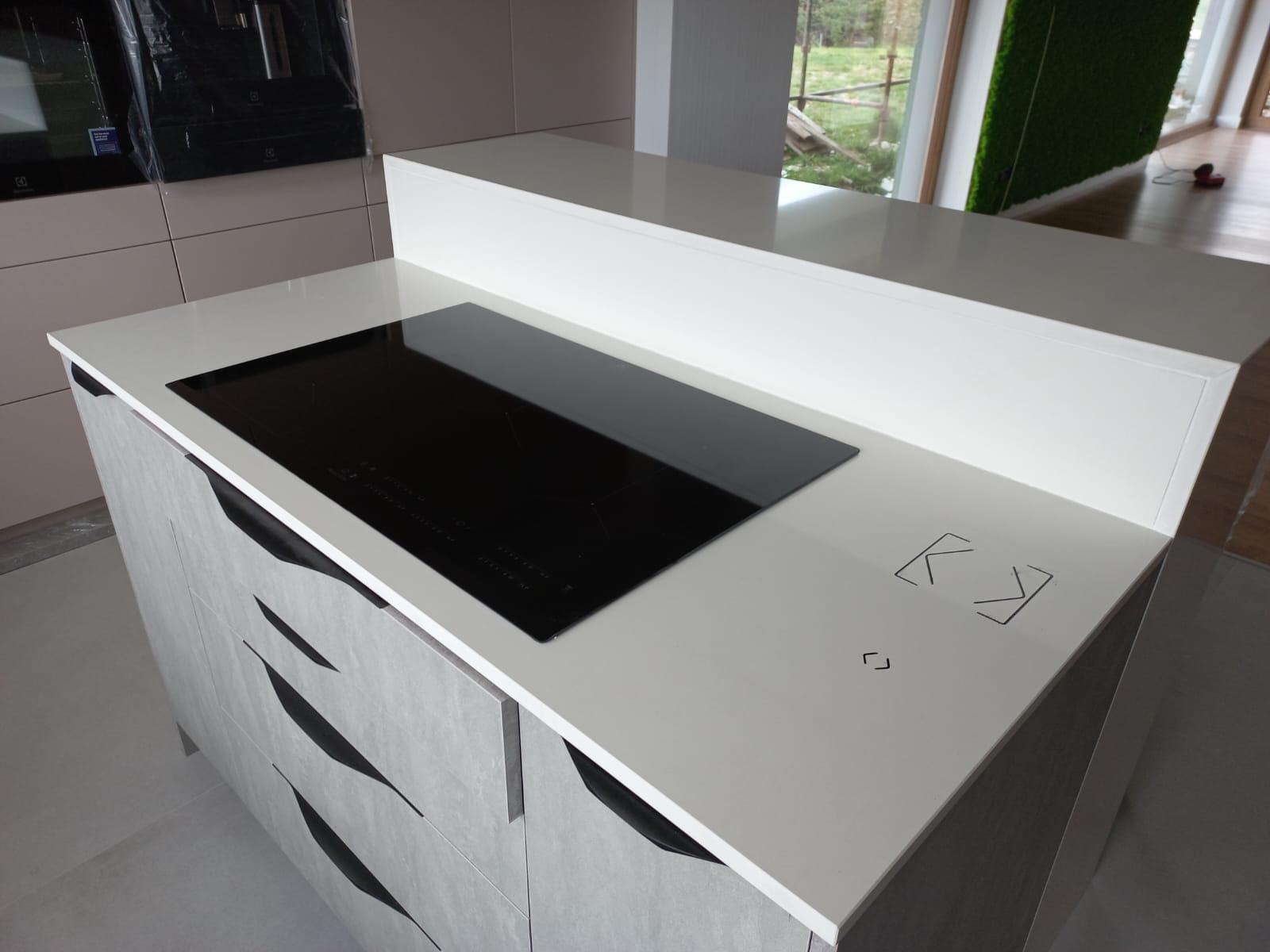 Biały blat kuchenny z wbudowaną czarną płytą indukcyjną i minimalistycznymi symbolami sterowania dotykowego, szare szafki z charakterystycznymi uchwytami, widok z boku.