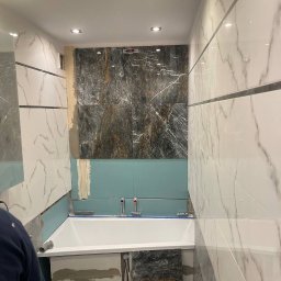 TamamBud - Biała wanna zabudowana płytami gipsowo-kartonowymi, częściowo wyłożona marmurowymi płytkami w odcieniach szarości i bieli, widoczne narzędzia glazurnicze i wiadro z zaprawą.