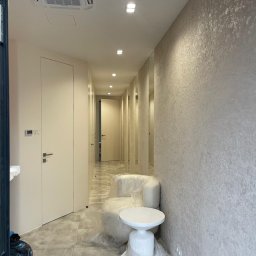 TamamBud - Korytarz w jasnych odcieniach z wykończonymi ścianami o fakturze, zabezpieczone folią meble, widoczne drzwi do WC i klimatyzacja sufitowa.