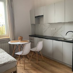 TamamBud - Wnętrze kawalerki z aneksem kuchennym: białe szafki kuchenne z czarnym blatem i marmurowym backsplashiem, okrągły stół z dwoma krzesłami, łóżko z wzorzystą pościelą, drewniana podłoga, roślina...