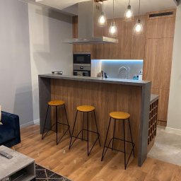 TamamBud - Nowoczesna kuchnia z wyspą i trzema hokerami, drewniane fronty szafek, szary blat, oświetlenie wiszące z czterema żarówkami w metalowych kloszach, drewniana podłoga i dywan w salonie.