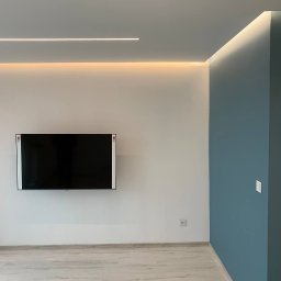 TamamBud - Minimalistyczne wnętrze po remoncie: białe ściany, jedna ściana pomalowana na niebiesko, podwieszony telewizor, panele podłogowe w odcieniach szarości i białe listwy przypodłogowe, oświetlenie LED...