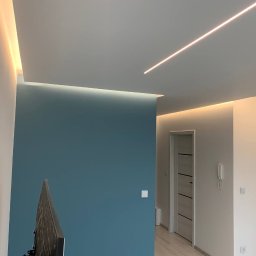 TamamBud - Nowoczesne wnętrze po odświeżeniu: błękitna ściana kontrastuje z białymi ścianami i sufitem z liniowym oświetleniem LED; widoczna drabina malarska w rogu.