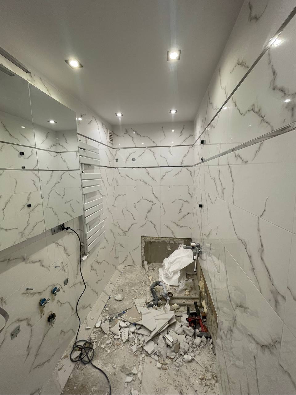 Wnętrze łazienki w trakcie prac: białe płytki imitujące marmur na ścianach, widoczne fragmenty instalacji, gruz i narzędzia na podłodze, brakujące płytki przyłączeniowe.