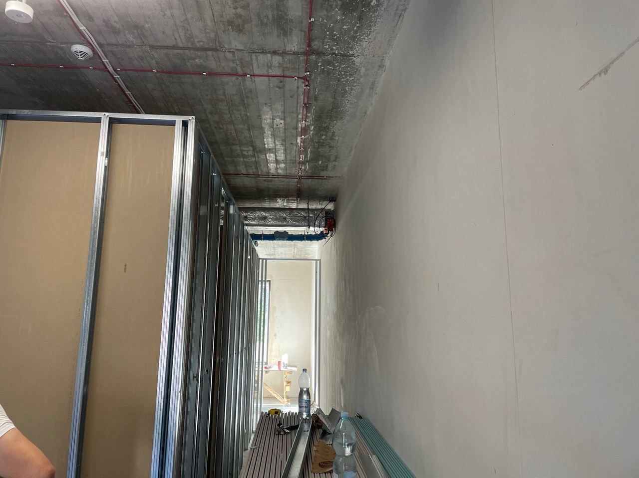 Wnętrze w trakcie prac: metalowy szkielet pod płyty karton-gips, surowy betonowy sufit z instalacją elektryczną, ściana po wstępnym nałożeniu gładzi, widoczne narzędzia i butelki wody.