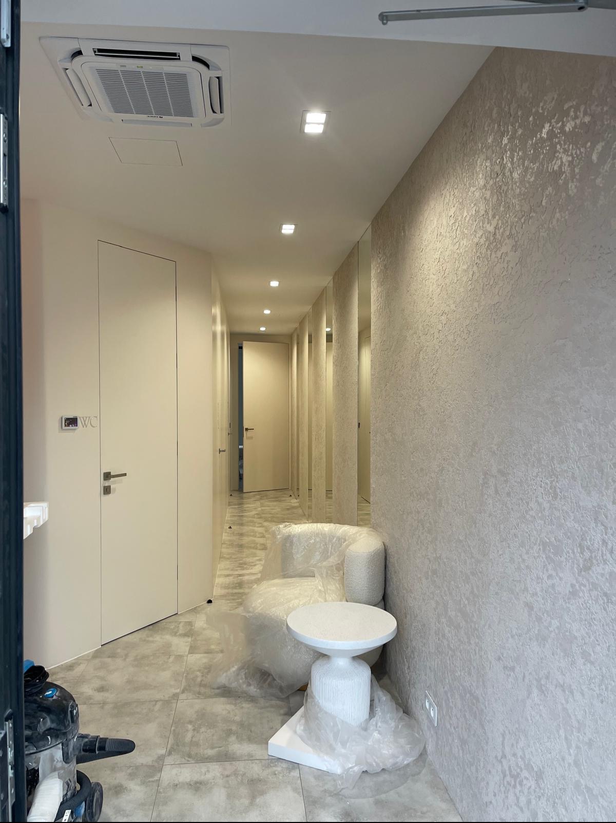 Korytarz w jasnych odcieniach z wykończonymi ścianami o fakturze, zabezpieczone folią meble, widoczne drzwi do WC i klimatyzacja sufitowa.