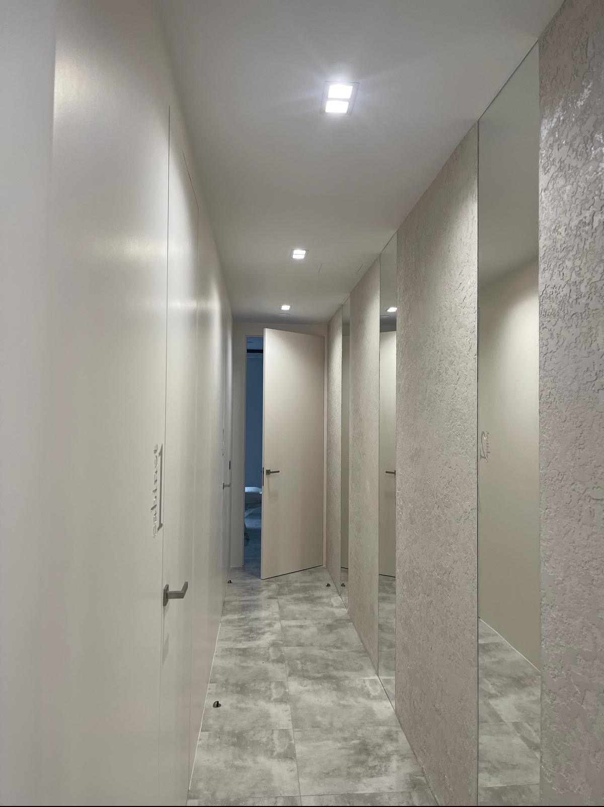 Nowoczesny korytarz z szarymi płytkami podłogowymi, jasnymi ścianami z dekoracyjną fakturą, wbudowanymi szafami z minimalistycznymi uchwytami i lustrami w wąskich panelach.