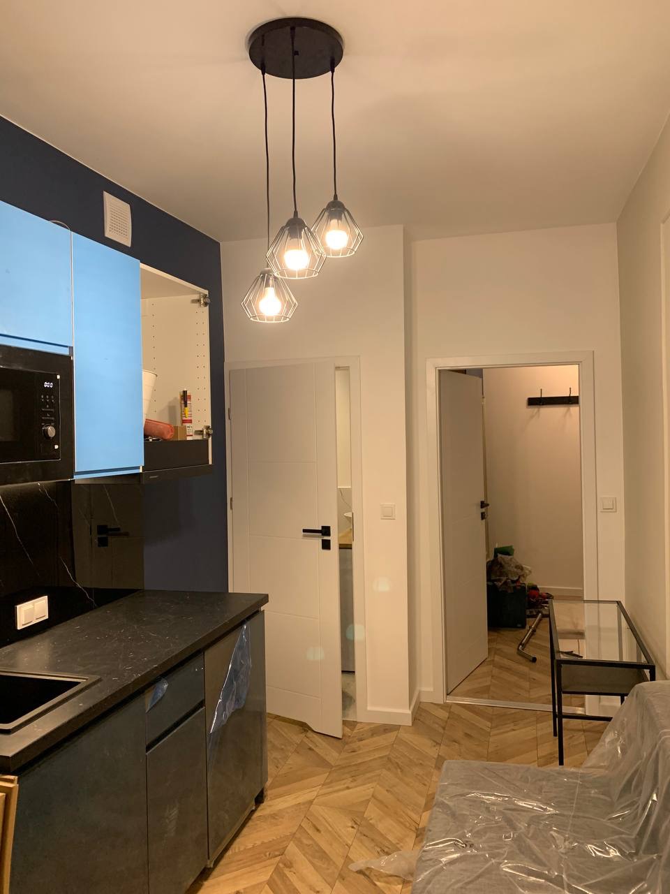 Nowoczesna kuchnia z czarnym blatem, niebieskimi szafkami i trzema wiszącymi lampami w geometrycznych kloszach, widok na parkiet ułożony w jodełkę i otwarte drzwi do kolejnego pomieszczenia.