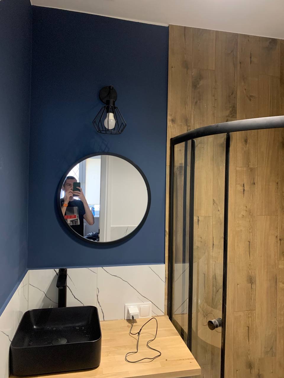 Nowoczesna łazienka w Warszawie z ciemnoniebieską ścianą, okrągłym lustrem z czarną ramą i geometryczną lampą. Umywalka nablatowa w kolorze czarnym, drewniany blat i kabina prysznicowa z czarnymi...