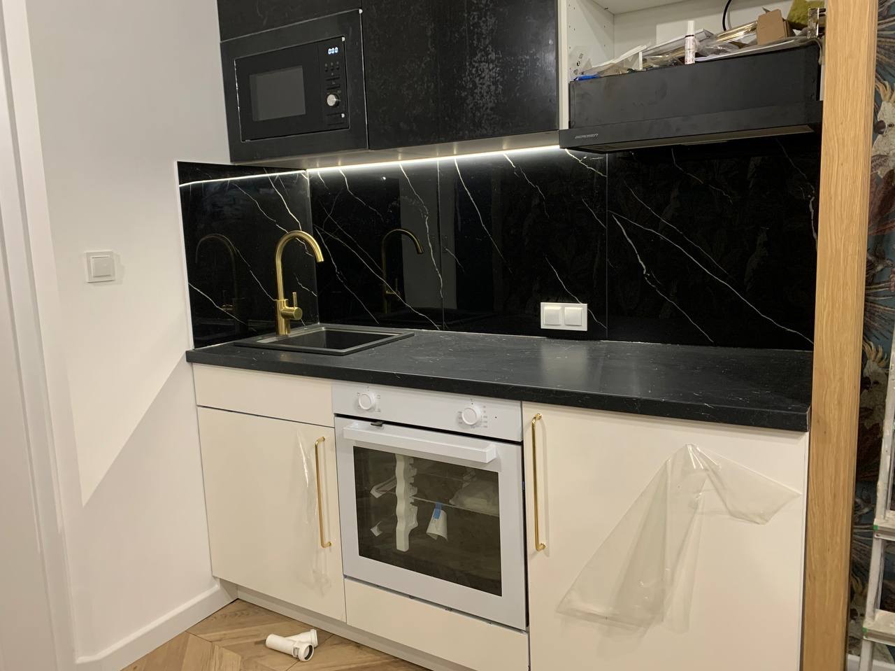 Częściowo wykończona kuchnia z czarnym blatem i panelem ściennym imitującym marmur, złotą armaturą i beżowymi szafkami, z widoczną rurą PCV na podłodze i folią ochronną na szafce.