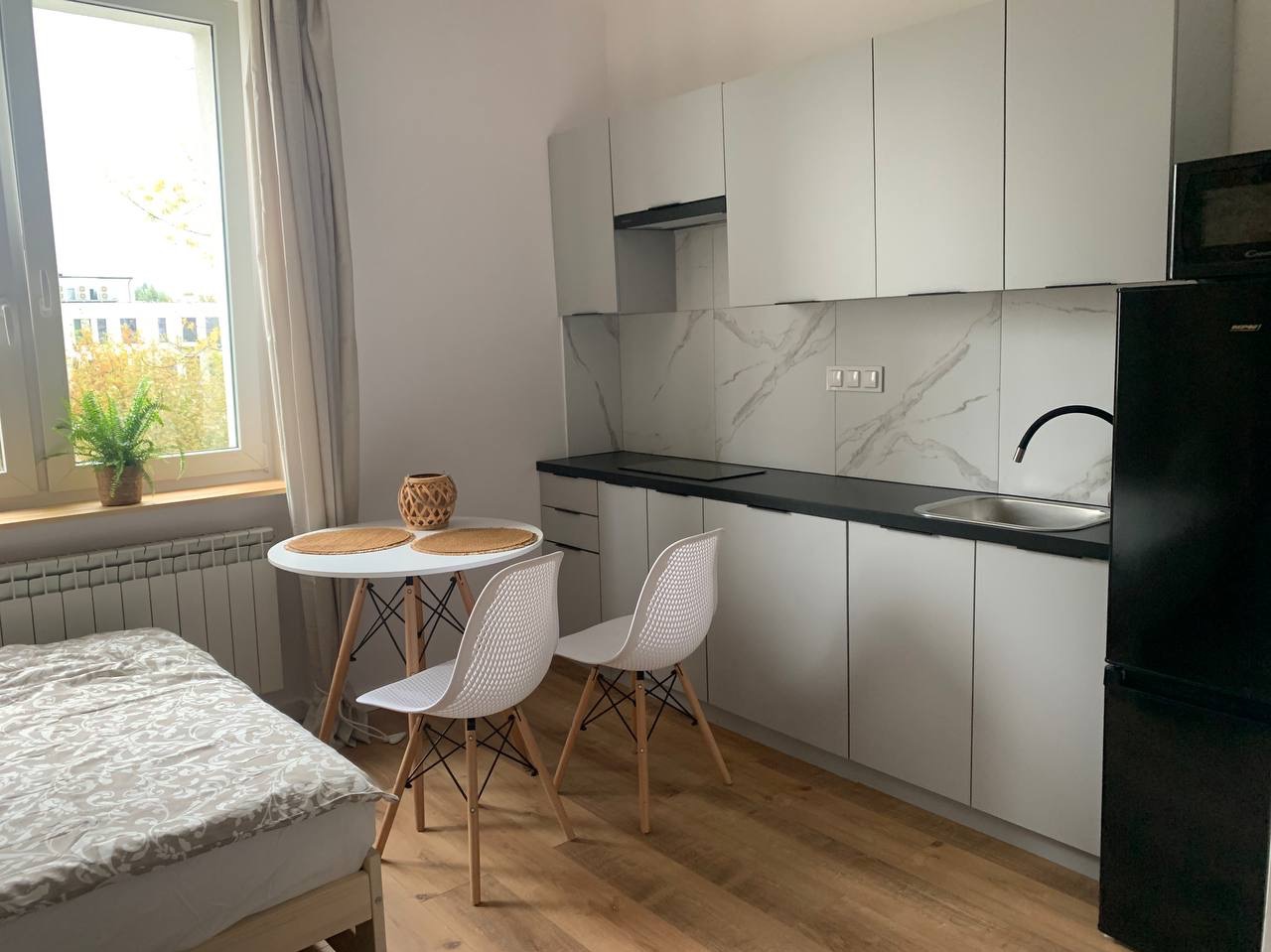 Wnętrze kawalerki z aneksem kuchennym: białe szafki kuchenne z czarnym blatem i marmurowym backsplashiem, okrągły stół z dwoma krzesłami, łóżko z wzorzystą pościelą, drewniana podłoga, roślina...