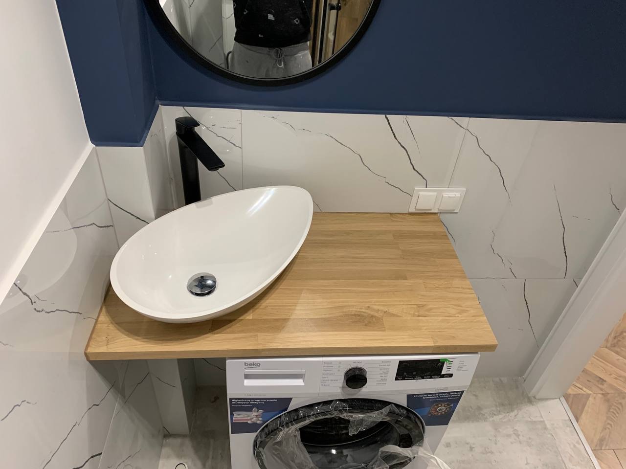 Nowoczesna łazienka z umywalką nablatową o owalnym kształcie i czarną armaturą, zamontowaną na drewnianym blacie nad pralką Beko, białe płytki z szarymi żyłkami na ścianie, widoczne odbicie osoby...