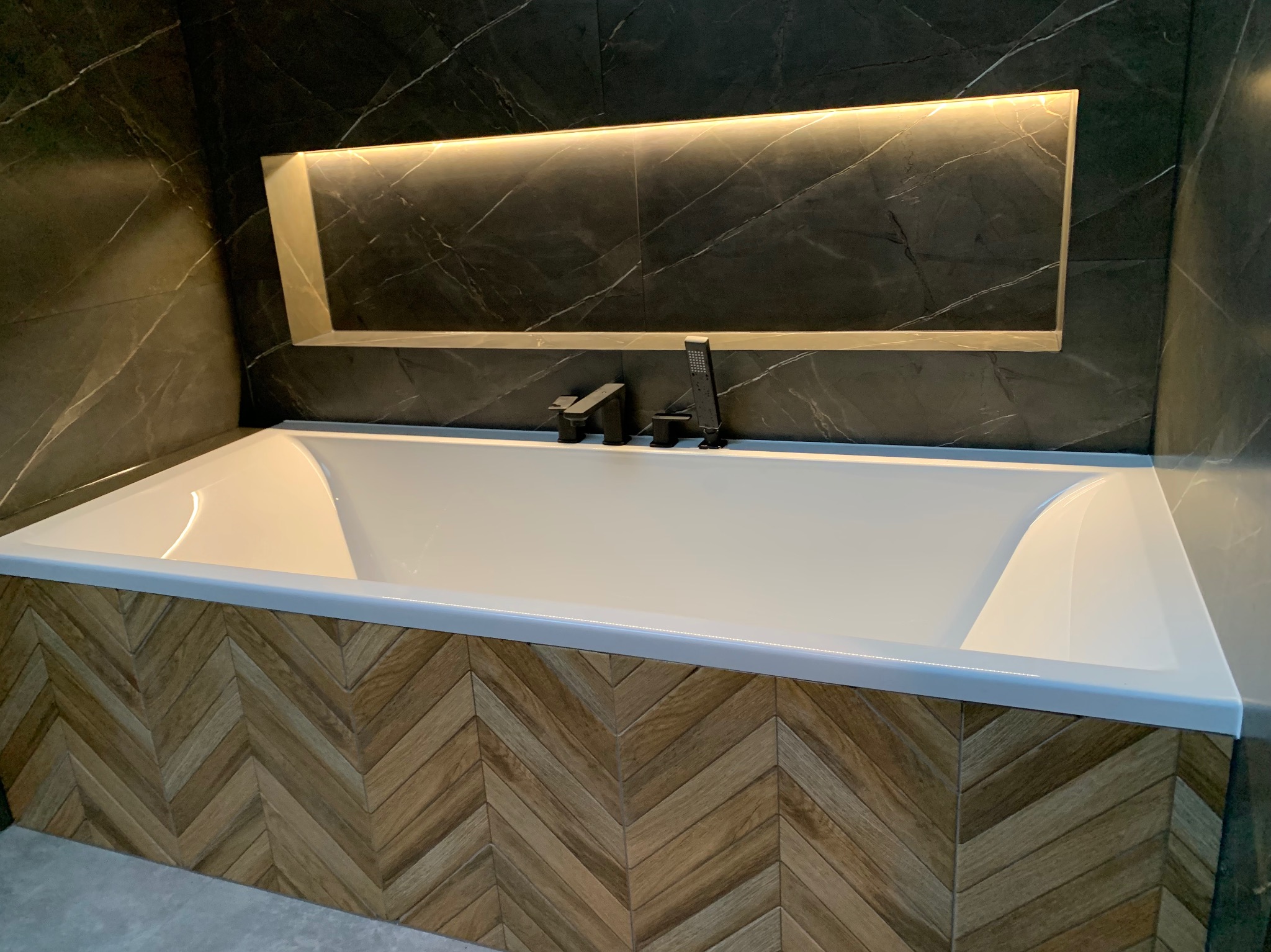 Nowoczesna łazienka z białą wanną obudowaną drewnopodobnymi panelami ułożonymi w jodełkę, czarna armatura i podświetlane wnęki w ciemnych, marmurkowych płytkach.