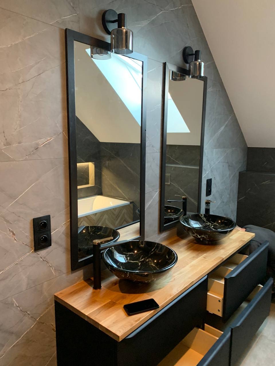 Nowoczesna łazienka z dwoma czarnymi umywalkami z marmurkowym wzorem, osadzonymi na drewnianym blacie szafki z czarnymi frontami, z dwoma wysokimi, prostokątnymi lustrami w czarnych ramach...