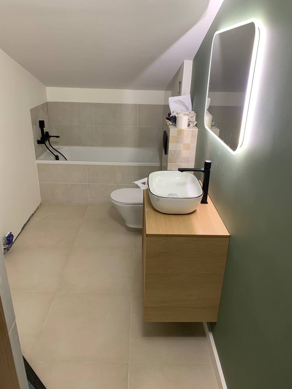 Nowoczesna łazienka w trakcie wykańczania: beżowe płytki, biała wanna z czarną armaturą, wisząca szafka z umywalką nablatową i podświetlane lustro.