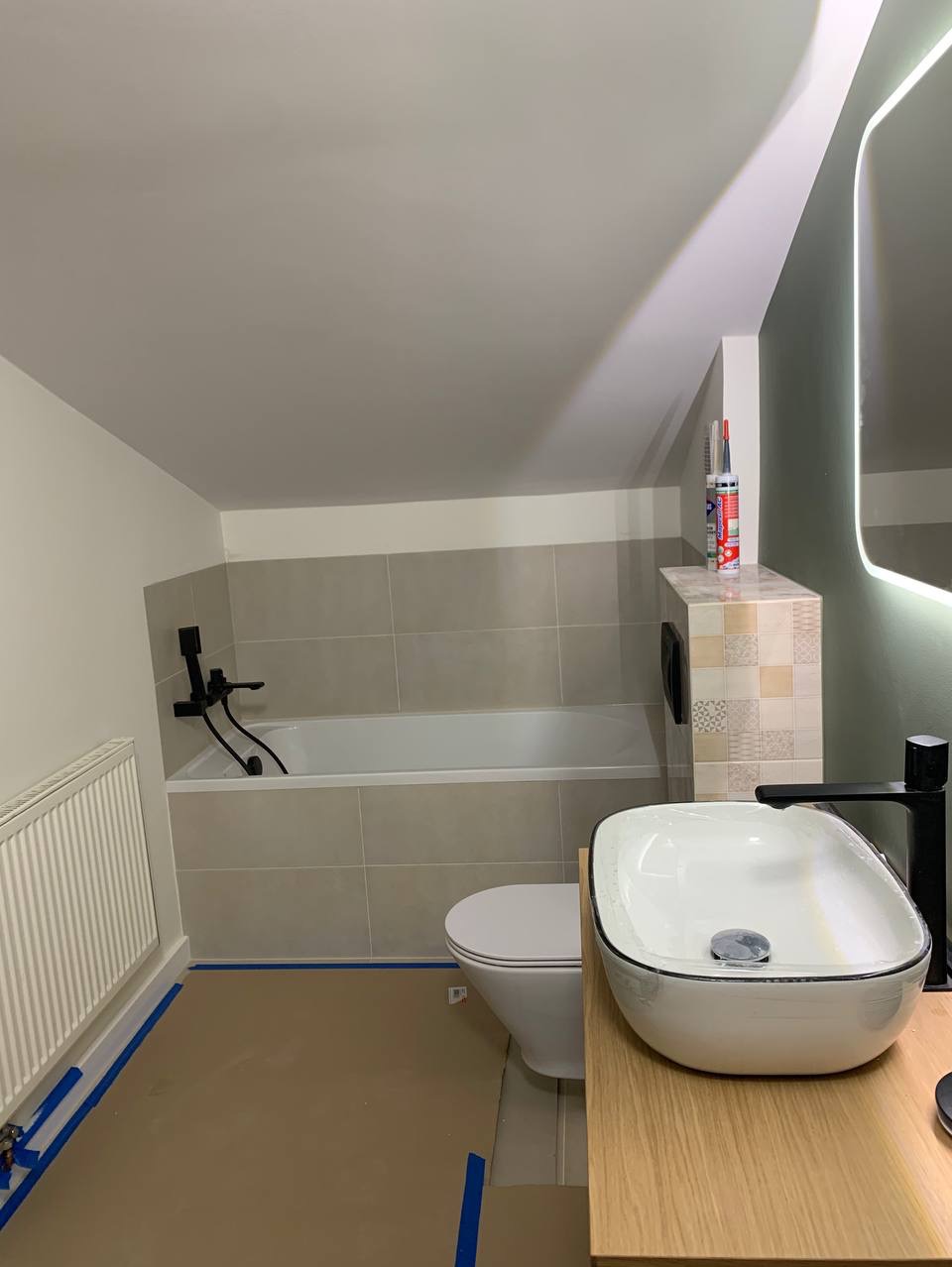Łazienka na poddaszu w trakcie prac wykończeniowych: zabezpieczona podłoga, biała wanna z czarną armaturą, umywalka nablatowa na drewnianym blacie, toaleta i częściowo wykończone płytki na ścianach.