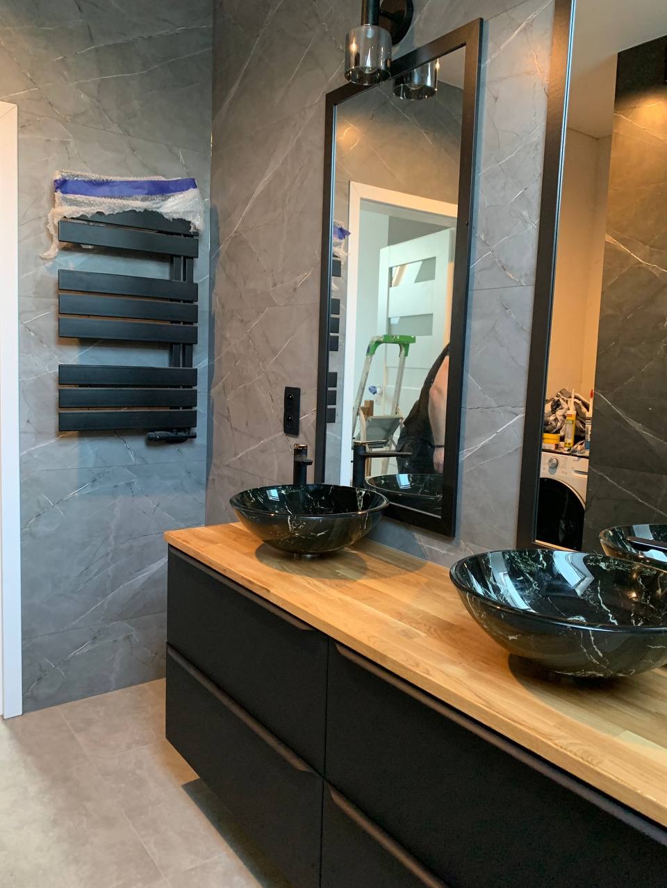 Nowoczesna łazienka z marmurowymi płytkami, czarną szafką z drewnianym blatem, dwoma umywalkami w kolorze czarnego marmuru oraz czarnym grzejnikiem drabinkowym w folii ochronnej.